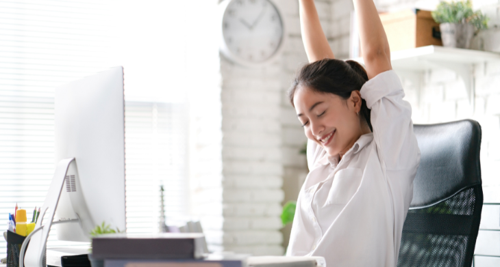 Das Bild zeigt eine Frau, die an ihrem Arbeitsplatz die Arme nach oben streckt und lächelt. Sie sitzt auf einem ergonomischen Bürostuhl vor einem Schreibtisch mit einem Monitor, Büchern und Büromaterial. Im Hintergrund sind ein großes Fenster, eine Uhr und Pflanzen zu sehen. Das Bild betont die Bedeutung von Bewegung und kurzen Pausen für mehr Wohlbefinden am Arbeitsplatz