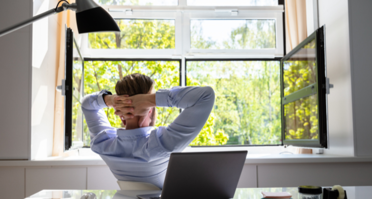 Mann in Businesskleidung, der entspannt auf einem Stuhl sitzt und aus dem Fenster blickt. Das Fenster ist weit geöffnet, sodass frische Luft und Tageslicht in den Raum strömen. Ein Laptop steht auf dem Schreibtisch, zusammen mit Büroartikeln. 