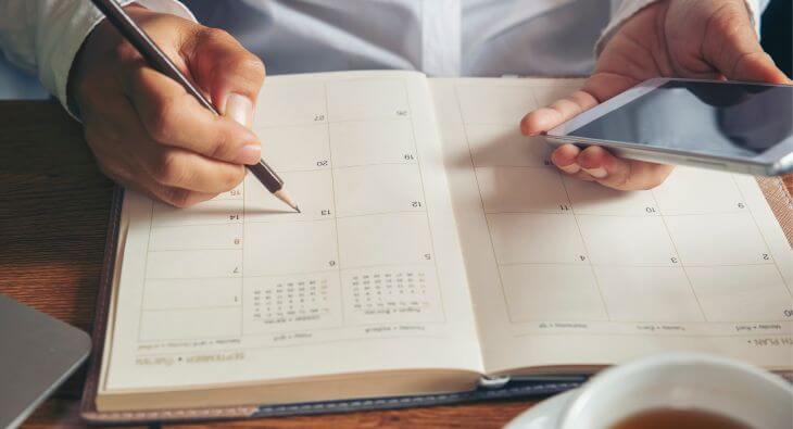 Ein Buchkalender liegt auf einem Tisch. Jemand trägt mit einem Bleistift etwas in den Terminkalender ein und hält in der anderen Hand ein Smartphone. Im unteren Bildbereich steht in einem Textbalken „Alles rund um Kalender für Büro und zu Hause – Welcher Kalender passt zu Ihnen?“