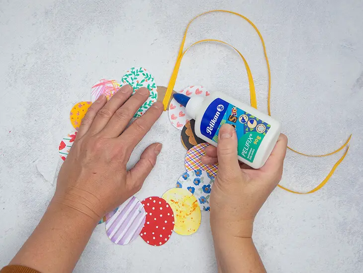 Jemand klebt mit Bastelkleber von Pelikan ein Textil-Geschenkband als Aufhänger an einen selbstgebastelten Osterkranz aus Papier. Foto: Johanna Rundel