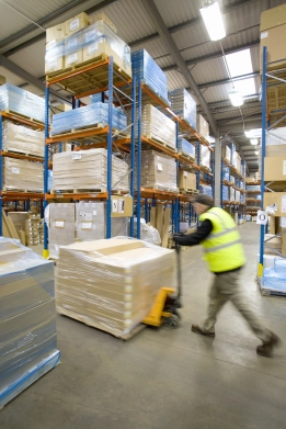 Lagerarbeiter mit Warnweste transportiert Ware mit Hubwagen in einem Hochregallager.