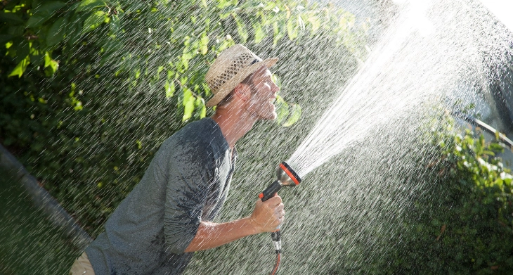 Mann mit Strohhut spritzt sich mit einer Gardena Gartenbrause nass, während er im Garten steht.
