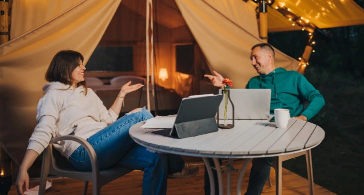 Zwei Personen arbeiten entspannt mit Laptops vor Zelt im Abendlicht.