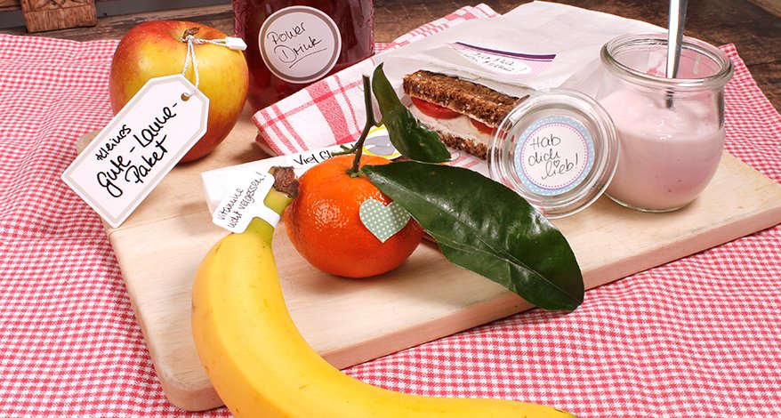 Auf einer rot-weiß karierten Tischdecke liegt ein Holzbrettchen. Darauf liegen eine Banane, eine Mandarine, ein Apfel, der ein Etikett der Marke Avery Zweckform angehängt hat mit der Aufschrift "Kleines Gute-Laune Paket". Auf dem Brettchen steht außerdem ein kleines Weckglas, welches einen Etiketten-Aufkleber mit der Aufschrift "Hab dich lieb!" Eine Butterbrot-Tüte mit einem Brot ist auch mit einem Etikett beklebt.