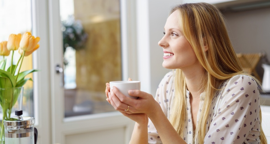 Eine Frau sitzt mit einer Tasse Kaffee in der Hand vor einem Fenster und schaut nach draußen. Auf dem Tisch vor ihr steht ein Strauß gelber Tulpen und eine Kanne Kaffee. 