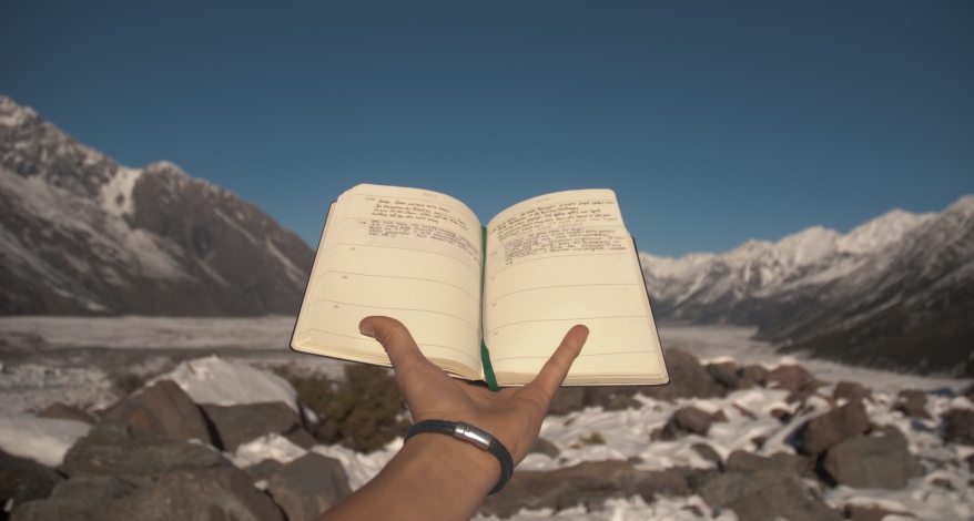 Eine Hand hält einen geöffneten Kalender von sich weg. Im Hintergrund ist eine Berglandschaft mit schneebedeckten Bergen zu sehen.