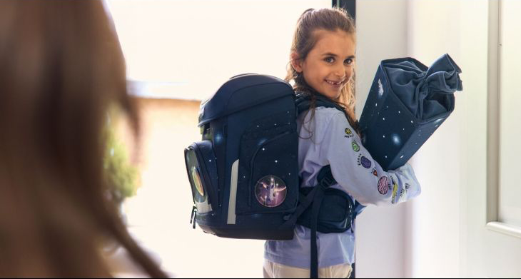 Ein Mädchen mit Schulranzen und Schultüte von ergobag steht im Hausflur und dreht sich zurück zu ihrer Mutter