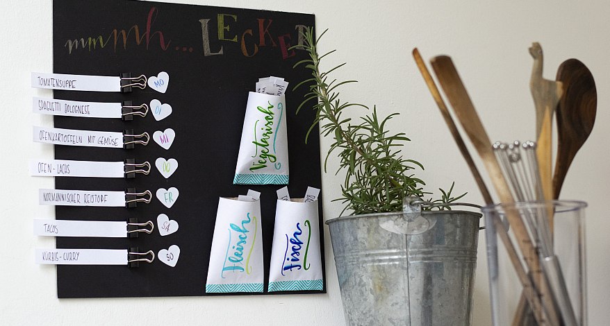 Selbstgebastelter Wochenplan mit Rezeptideen auf schwarzem Brett neben Kochutensilien und Rosmarin.