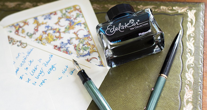 Auf einer Schreibunterlage liegt ein offener Briefumschlag mit einen handschriftlichen Brief. Daneben liegt ein Pelikan Füller, ein Pelikan Kugelschreiber und eine Edelstein Tinte von Pelikan.