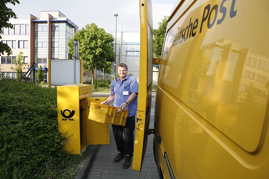 Ein Breifträger leert einen Briefkasten und trägt die Kiste mit der Post in das Postauto.