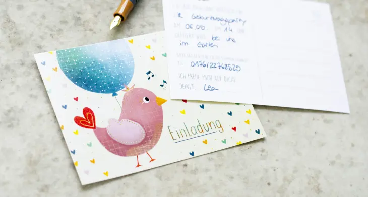 Eine Karte, die einen Vogel, einen Luftballon sowie das Wort "Einladung" zeigt, liegen auf einem Betontisch. Eine beschriebene Postkarte und ein Füller liegen ebenfalls darauf.