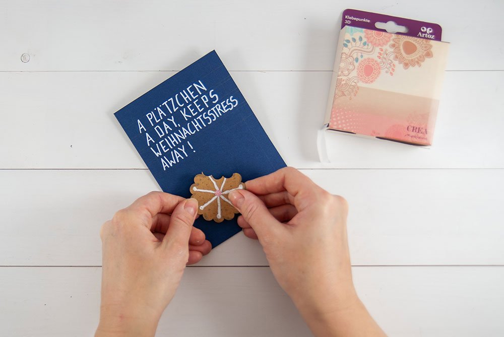 Auf einem weißen Holzuntergrund liegt eine blaue Karte, die mit dem Text "A Plätzchen a day, keeps the Weihnachtsstress away!" beschriftet wurde. Darauf kleben zwei Hände einen braunen Motivkarton, der wie ein Plätzchen geformt und bemalt ist. Am rechten, oberen Bildrand lieben Klebepunkte der Marke Artoz.