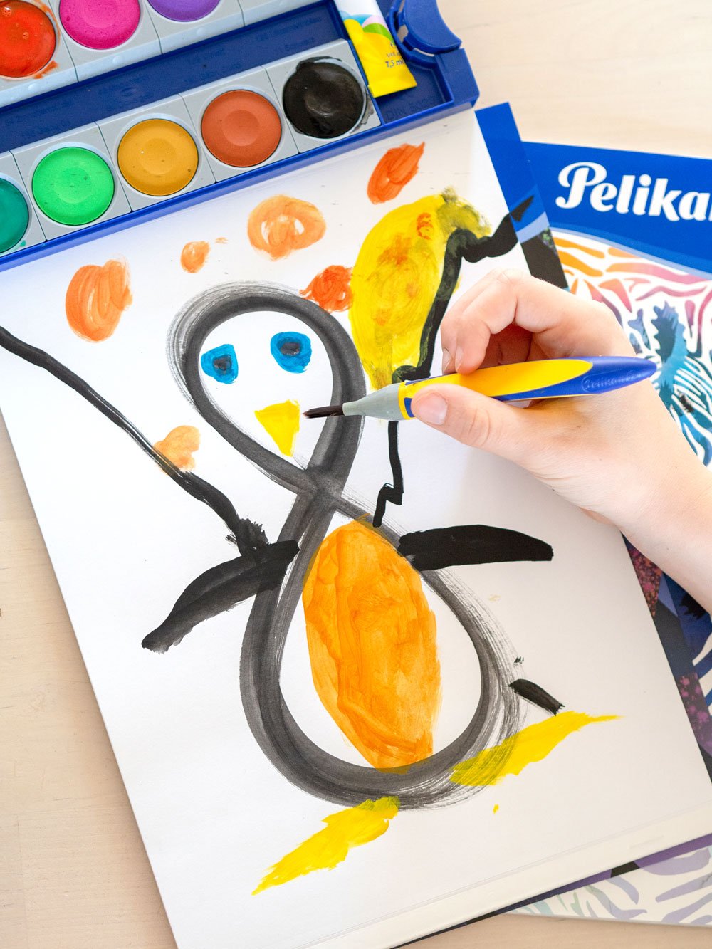 Kinderhand malt mit Wasserfarben aus einer 8 einen bunten Pinguin, daneben Wasserkasten