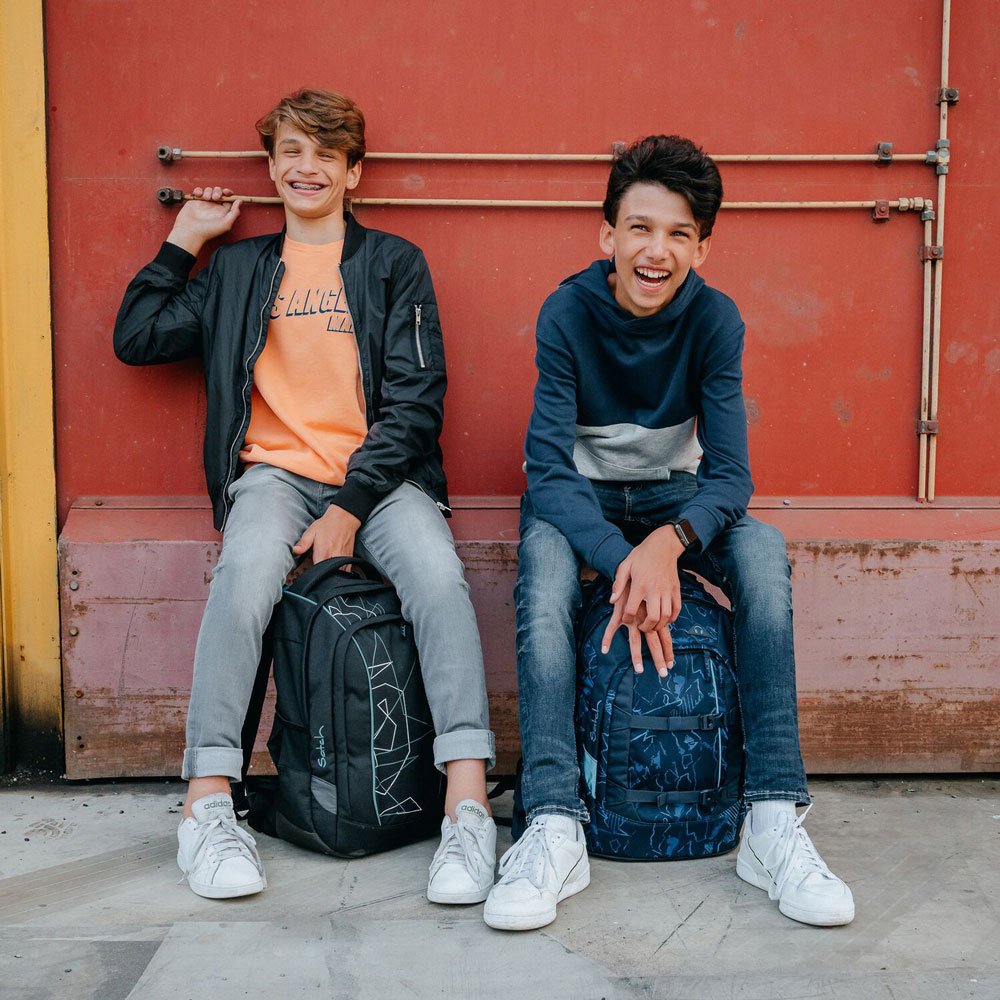 Zwei Jungen sitzen auf einem Industriegelände auf einem Vorsprung und lachen. Sie haben beide einen Rucksack der Marke satch dabei.