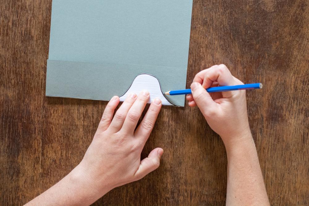 Ein blaues Stück Papier liegt auf einem Holztisch. Eine Hand hält die Schablone an der Falz fest, die andere Hand fährt mit einem Belistift um die Schablone.