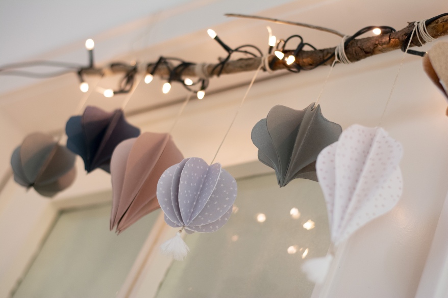Eine aus Papier gebastelte Girlade als Tropfen und Kugeln in unterschiedlichen Farben hängen an einem beleuchteten Ast vor einem Fenster.