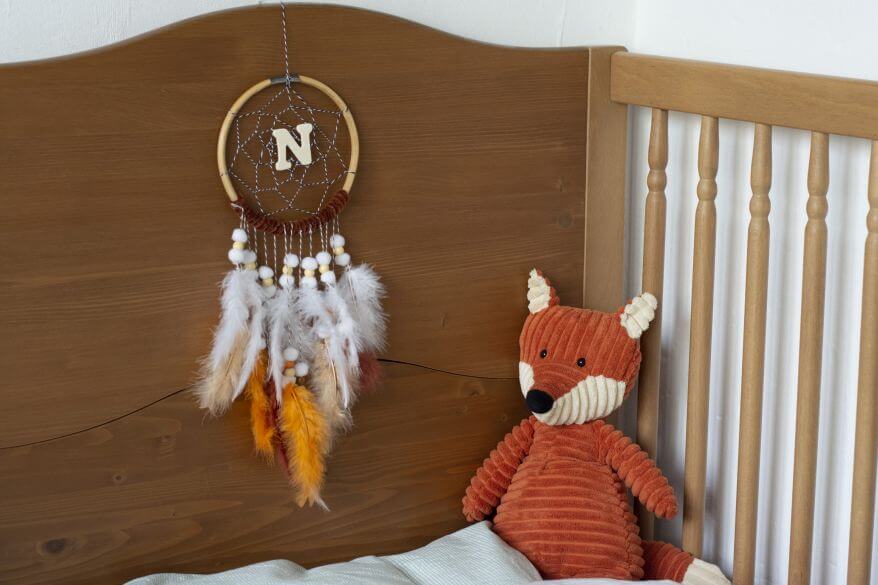 Ein selbstgebastelter Traumfänger mit dem Buchstaben N in der Mitte hängt über einem Babybett, daneben sitzt ein Fuchs als Kuscheltier.