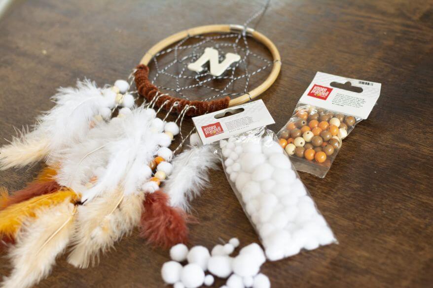 Blick auf einen selbstgebastelten Traumfänger, der auf einem Tisch liegt. Daneben liegen Pompons und Holzperlen von Knorr Prandell.