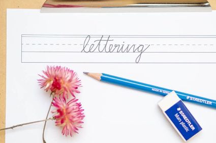 Auf einem Klemmbrett aus Holz klemmt ein Papier mit Hilfslinien zum Lettern. In den Zeilen steht das Wort „lettering“ in klassischem Handlettering-Stil. Bleistift und Radiergummi der Firma Staedtler liegen darunter. Zwei rosa Trockenblumen lockern das Bild auf. Foto: Johanna Rundel