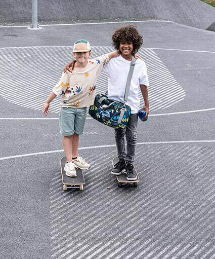 Zwei Grundschüler stehen auf ihren Skateboards auf dem Schulhof und halten sich aneinander fest. Der Junge auf der rechten Seite hat dabei eine Sporttasche der Marke School-Mood umgehängt und hält eine Trinkflasche in der Hand.