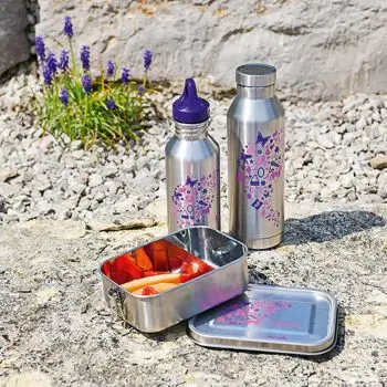 Zwei Trinkflaschen und eine Brotdose aus Metall der Marke Step-by-Step sind im Freien in einem Steingarten aufgestellt. Die Brotdose ist geöffnet, man sieht rote Paprikastreifen.