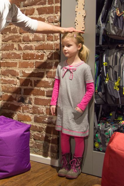 Ein Mitarbeiter eines Schulranzen-Fachgeschäfts ermittelt anhand einer Skala an der Wand die Körpergröße einer zukünftigen Erstklässlerin.