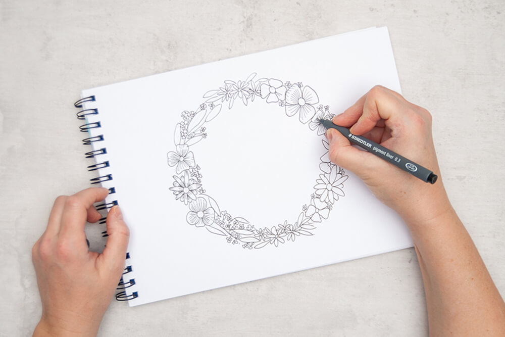 Jemand zeichnet mit einem schwarzen Pigment Liner von Staedtler Blüten und Blätter entlang eines Bleistift-Kreises auf einem Malblock. Foto: Johanna Rundel