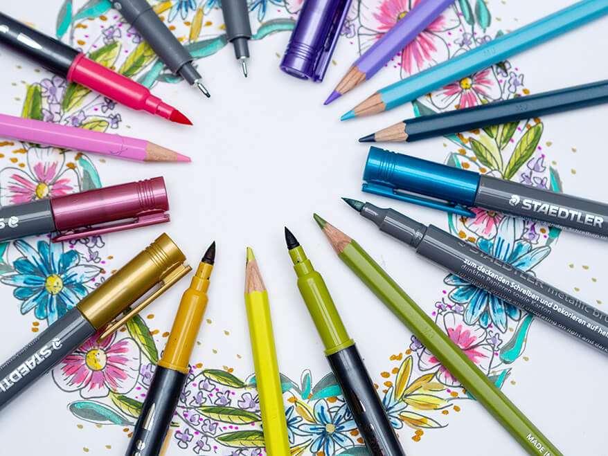 Verschiedene Farbstifte sowie Pigment Liner der Marke Staedtler liegen im Kreis auf der Zeichnung eines Blumenkranzes. Foto: Johanna Rundel