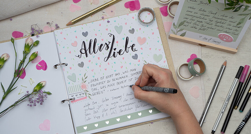 Auf einem hellen Untergrund liegt ein aufgeschlagenes Hochzeitsgästebuch von Goldbuch. In großen Letteringbuchstaben steht dort „Alles Liebe“ geschrieben. Jemand schreibt gerade Glückwünsche hinein. Die Seite ist kreativ mit Aufklebern, Washi Tape und bunten Stiften gestaltet. Die benutzen Materialien liegen um das Buch verstreut. In der rechten oberen Ecke ist ein Holzaufsteller der Marke Goldbuch mit einer Anleitungskarte zu sehen. Überall sind Papierherzen und Blümchen verstreut. Foto: Johanna Rundel