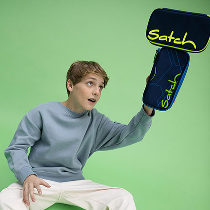 Vor einem hellgrünen Hintergrund balanciert ein Jugendlicher in beiger Hose und taubenblauem Pullover zwei Schlamperboxen von satch in einer Hand.