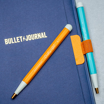 Auf einem hellgrauen Untergrund liegt ein dunkelblaues Bullet Journal von Leuchtturm1917. Darauf liegt ein orangefarbener Drehgriffel. Ein türkiser Drehgriffel steckt in einer orangenen Stiftschlaufe, die am Journal befestigt ist. Foto: Johanna Rundel 