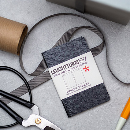 Auf einem hellgrauen Untergrund liegt ein schwarzer Geburtstagskalender im Taschenformat von Leuchtturm1917. Drumherum sind verschiedenen Gegenstände zum Geschenkeeinpacken verteilt: eine schwarze Metallschere, eine Rolle Packpapier, dunkelgraues Geschenkband und ein orangener Drehgriffel. Foto: Johanna Rundel 