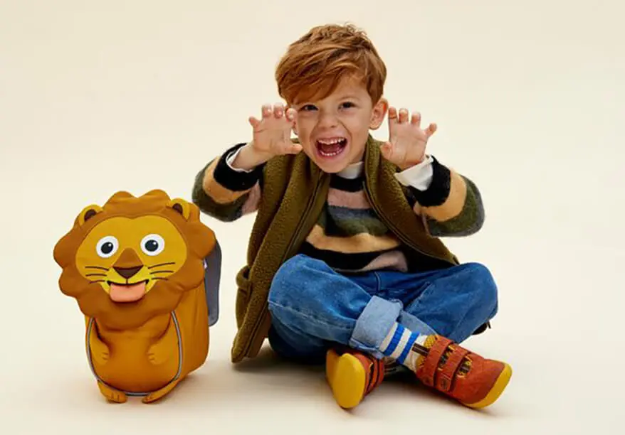 Ein Junge sitzt im Schneidersitz neben seinem Löwen-Rucksack von Affenzahn und brüllt mit zu Krallen gehobenen Händen in die Kamera.