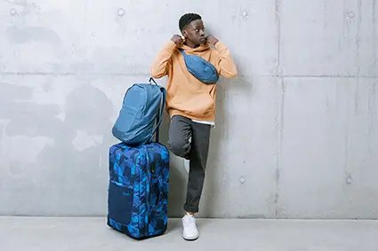Ein Teenager steht mit seinem Reisegepäck von satch lässig an eine Betonwand gelehnt und zieht sich gerade den Gurt eines blauen Hip-Bag über den Kopf.