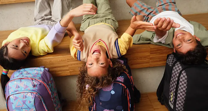 Drei Schüler liegen kopfüber auf einer Holztreppe auf ihren satch-Schulrucksäcken und lachen in die Kamera.