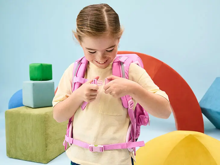 Eine Grundschülerin im hellgelben T-Shirt befestigt den Brustgurt ihres rosafarbenen Schulranzens von ergobag.)