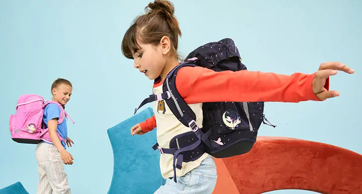 Zwei Grundschülerinnen toben mit Ihren Schulranzen von ergobag über verschiedene bunten Schaumstoffpolster. Der Hintergrund ist hellblau.