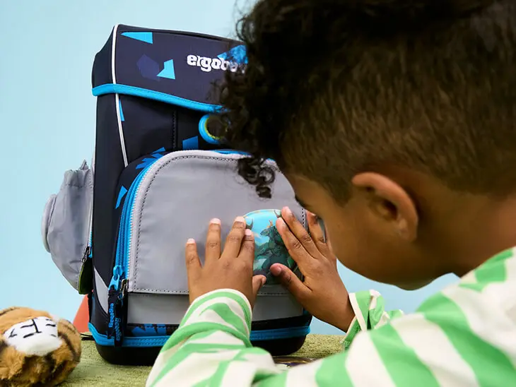 Ein dunkelhäutiger Junge im gestreiften Pullover befestigt ein Klettie an seinem blauen Schulranzen von ergobag.
