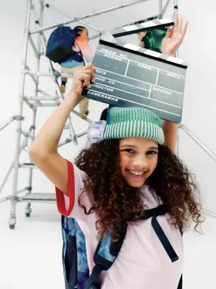 Ein Mädchen hält lächelnd eine Filmklappe über ihrem Kopf, im Hintergrund klettern Kinder auf einem Stahlgerüst.air auf ihrem Kopf.