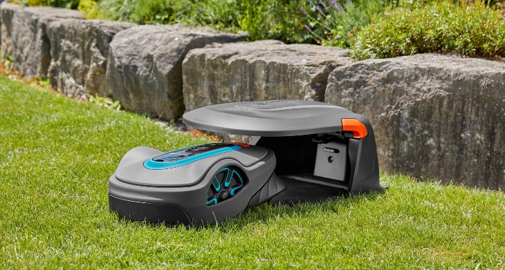 Gardena Mähroboter in einer Ladestation auf einer grünen Rasenfläche vor einer Natursteinmauer.