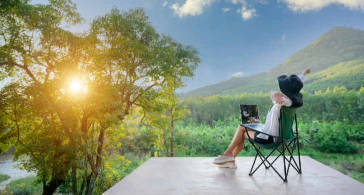 Frau im Outdoor-Homeoffice mit Laptop blickt in grüne Berglandschaft.