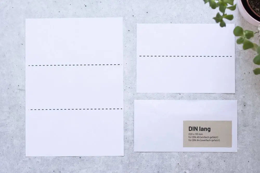 Auf grauem Grund liegt ein DIN A4-Papier mit zwei gestrichelten Markierungen, die das Papier in Drittel teilt und ein DIN A5-Papier mit einer gestrichelten Markierung in der Mitte, dazu ein passender Briefumschlag DIN lang. Auf dem Briefumschlag ist ein Aufkleber aufgebracht mit folgenden Infos: DIN lang, 220x110 Millimeter, für DIN A5 (einfach gefalzt), für DIN A4 (zweifach gefalzt). Rechts oben ragt der Teil einer Pflanze ins Bild.