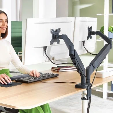 Das Bild zeigt eine Frau, die an einem Schreibtisch sitzt und an einem Computer arbeitet. Die beiden Monitore sind mithilfe von ergonomischen Monitorarmen befestigt und auf die richtige Höhe eingestellt. Die Frau wirkt entspannt und hat eine gesunde, aufrechte Sitzhaltung. 