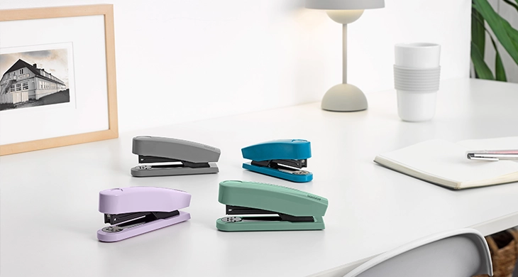 Das Bild zeigt vier farbige Novus-Heftergeräte in grau, türkis, lila, und mintgrün auf einem ordentlichen, minimalistischen Schreibtisch. Im Hintergrund sind eine Lampe, eine Tasse, ein Notizbuch und ein gerahmtes Foto zu sehen.