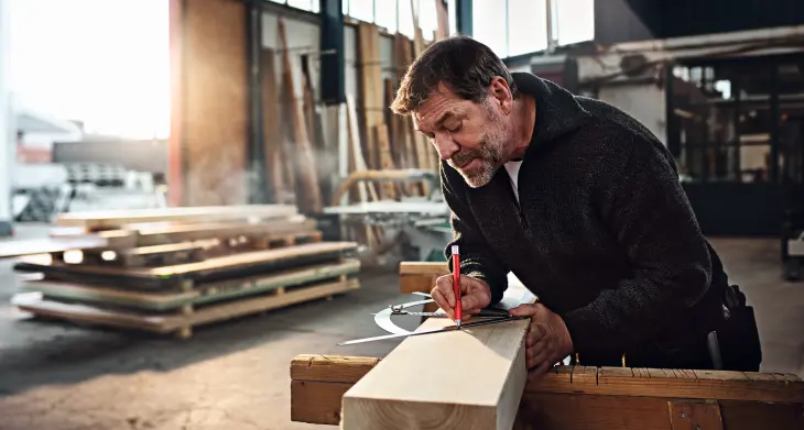Ein Mann mit Bart konzentriert sich, während er ein Stück Holz mit einem edding Druckbleistift abmisst und markiert. Er befindet sich in einer Tischlerwerkstatt, umgeben von verschiedenen Holzstücken und Werkzeugen.