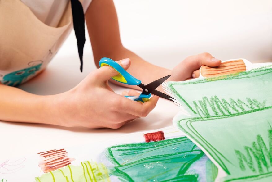 Nahaufnahme der Hand eines Kindes, die eine Schere der Serie Griffix von Pelikan hält. Die Klingen der Schere sind geöffnet, das Kind ist im Begriff, in ein bemaltes Papier zu schneiden.
