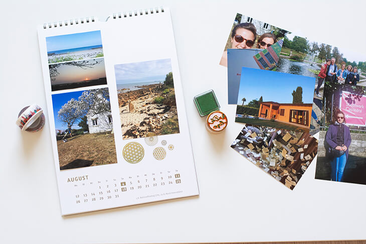 Auf weißem Untergrund liegt im linken Bildbereich ein Foto- und Bastelkalender, in dem sommerliche Urlaubsfotos eingeklebt sind. Rechts daneben liegen verschiedene Fotos auf einem Stapel, Washitape und ein Stempel mit Stempelkissen.