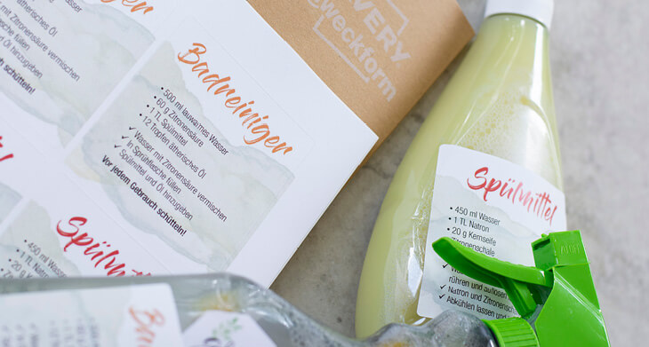 Rechts im Bild liegt eine Spülmittelflasche mit selbstgemachtem Spülmittel darin. Darauf klebt ein Etikett, auf dem „Spülmittel“ und die Anleitung steht. Links liegt ein Etikettenbogen von Avery Zweckform, auf dem Rezepte zu mehreren DIY-Haushaltsmitteln gedruckt sind. Am unteren Bildrand sieht man im Anschnitt eine Badreiniger-Flasche mit selbstgemachtem Badreiniger.