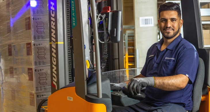 Lagerarbeiter fährt Gabelstapler mit Palette, trägt Arbeitskleidung und Sicherheitsausrüstung.