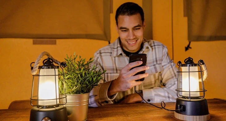 Mann im Zelt nutzt Handy, beleuchtet von zwei Campingleuchten.